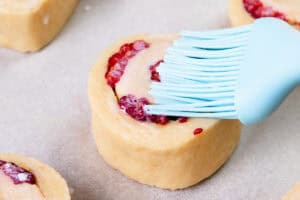 A close-up of a raw scone with raspberries on parchment paper being brushed with milk using a blue silicone pastry brush.