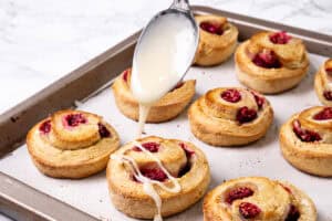 A spoon drizzling white glaze over freshly baked raspberry scones on a parchment-lined baking tray.