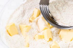 Cubed butter being blended into flour with a pastry cutter in a clear glass bowl.