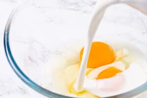 Cream being poured into a clear glass bowl with a whole egg, set on a marble countertop.