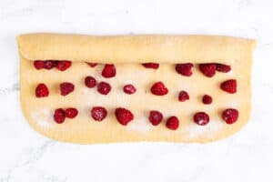 Scone dough on a marble surface, rolled halfway to enclose scattered fresh raspberries for filling.