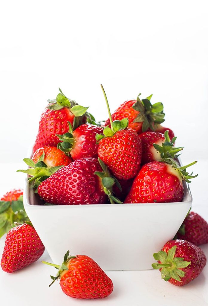 Fresh strawberries piled high in a bowl and laying next to it on the table