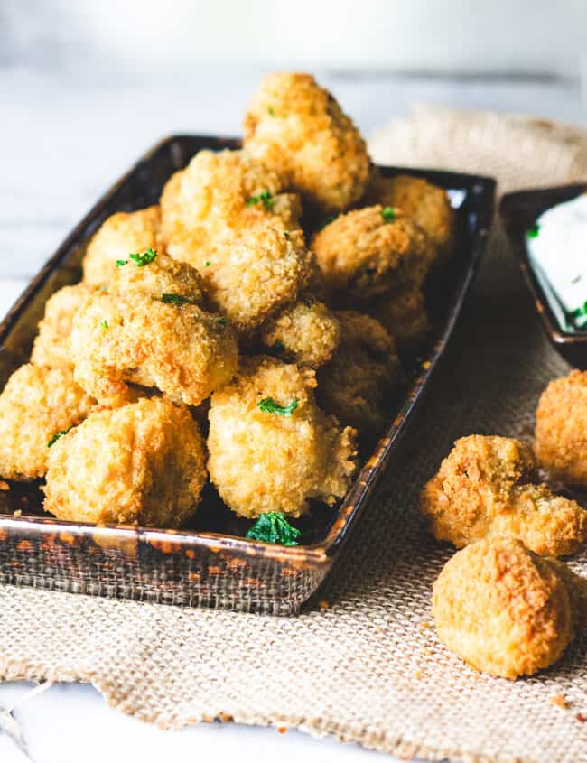 A close-up of a plate filled with golden brown, breaded mushrooms. One mushroom is being dipped into a white dipping sauce on the side.