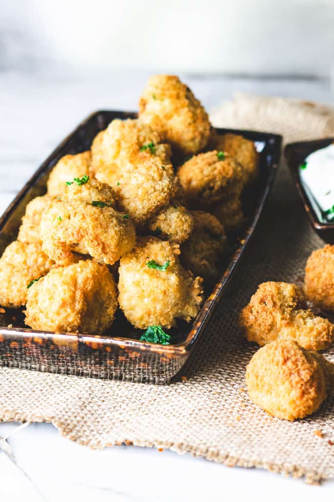 A close-up of a plate filled with golden brown, breaded mushrooms. One mushroom is being dipped into a white dipping sauce on the side.