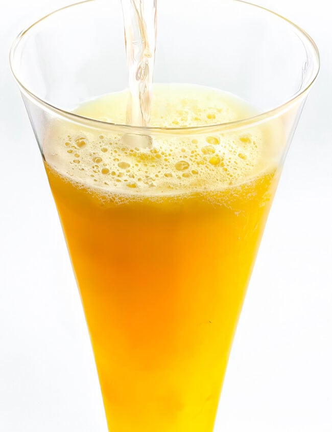A glass of Bellini cocktail being poured, with the fizzy peach-colored drink creating bubbles at the top.