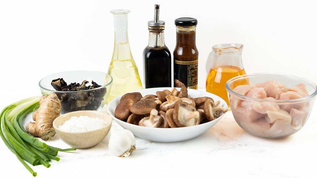 Various ingredients on a white surface for Chicken with Chinese Mushrooms: raw chicken pieces, shiitake mushrooms, dried mushrooms, ginger, garlic, green onions, cornstarch, oil, soy sauce, oyster sauce, and a glass container of honey.