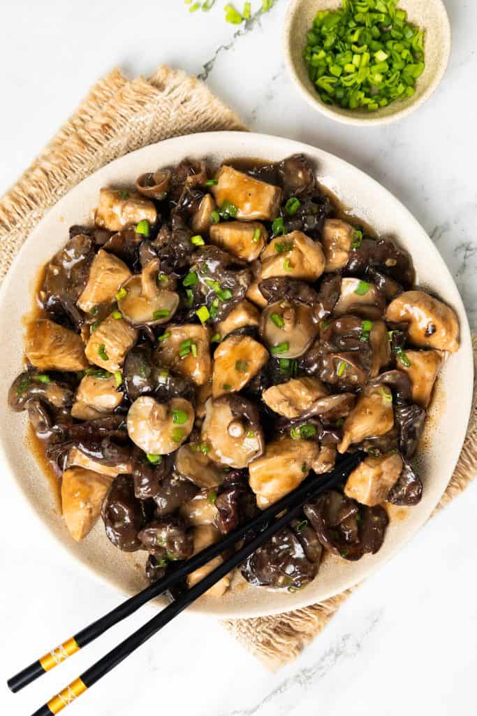 A plate of stir-fried chicken with Chinese mushrooms and green onions, served with black chopsticks, sits invitingly on a white surface. A small bowl of chopped green onions rests beside the plate on a woven mat, completing this flavorful presentation.