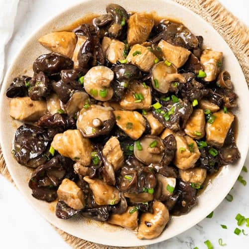 A plate of Chicken with Chinese Mushrooms in a dark sauce, garnished with chopped green onions, sits on a woven mat beside a bowl of rice, chopsticks, and a small bowl of extra green onions.