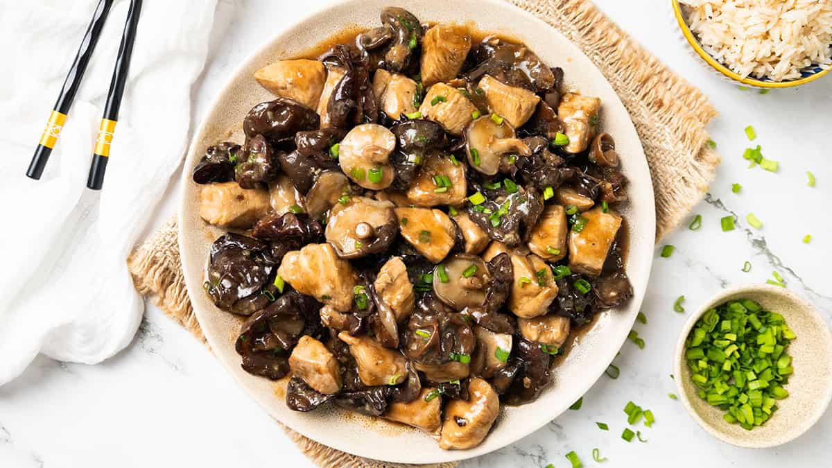 A plate of Chicken with Chinese Mushrooms in a dark sauce, garnished with chopped green onions, sits on a woven mat beside a bowl of rice, chopsticks, and a small bowl of extra green onions.