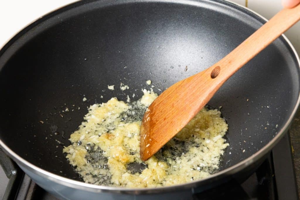 Minced garlic being sautéed in oil in a black pan, stirred with a wooden spatula over a stovetop—the perfect aromatic base for preparing Chicken with Chinese Mushrooms.