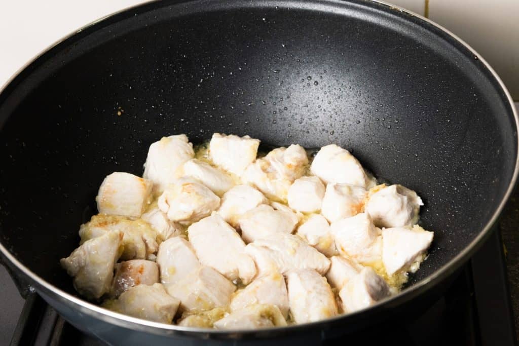 Chunks of chicken breast being sautéed in a black nonstick pan, lightly browned and cooking in oil on a stovetop, perfect for preparing classic Chicken with Chinese Mushrooms.