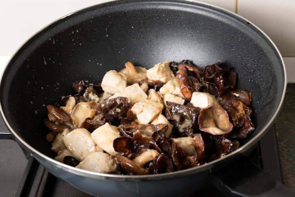 Chunks of chicken and wood ear mushrooms, reminiscent of the classic Chicken with Chinese Mushrooms, cooking in a black frying pan on a stovetop, with a white tiled wall in the background.