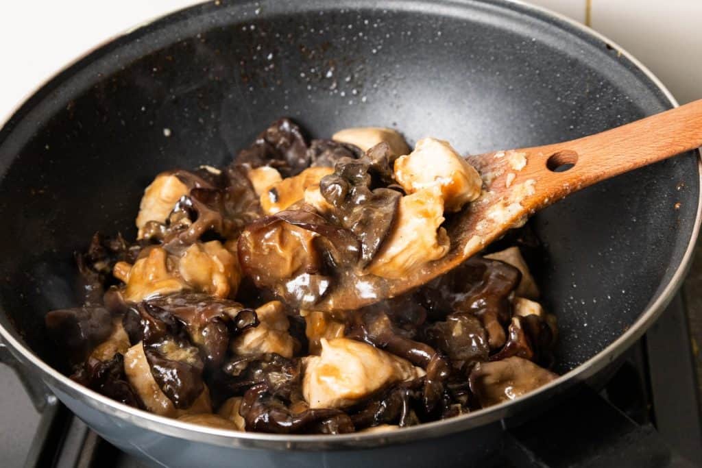 Chicken with Chinese Mushrooms and wood ear mushrooms being stir-fried in a black wok, with a wooden spatula stirring the ingredients and steam rising from the hot dish.