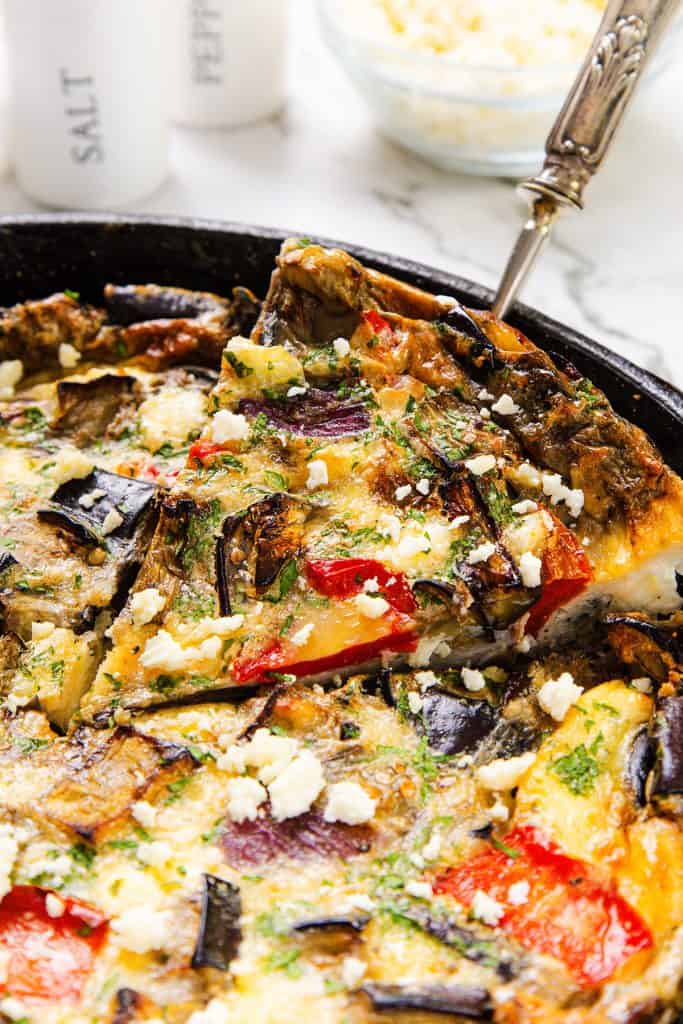 A close-up of a Baked Eggplant Frittata with Red Onion, Peppers & Feta in a skillet, filled with vibrant veggies, topped with herbs and crumbled cheese, as a serving is lifted by a spatula.