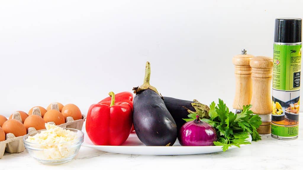 A plate with eggplant, red bell pepper, red onion, and parsley hints at a Baked Eggplant Frittata with Red Onion. Nearby are peppers & feta, eggs, grated cheese, cooking spray, and wooden salt and pepper grinders on a white surface.