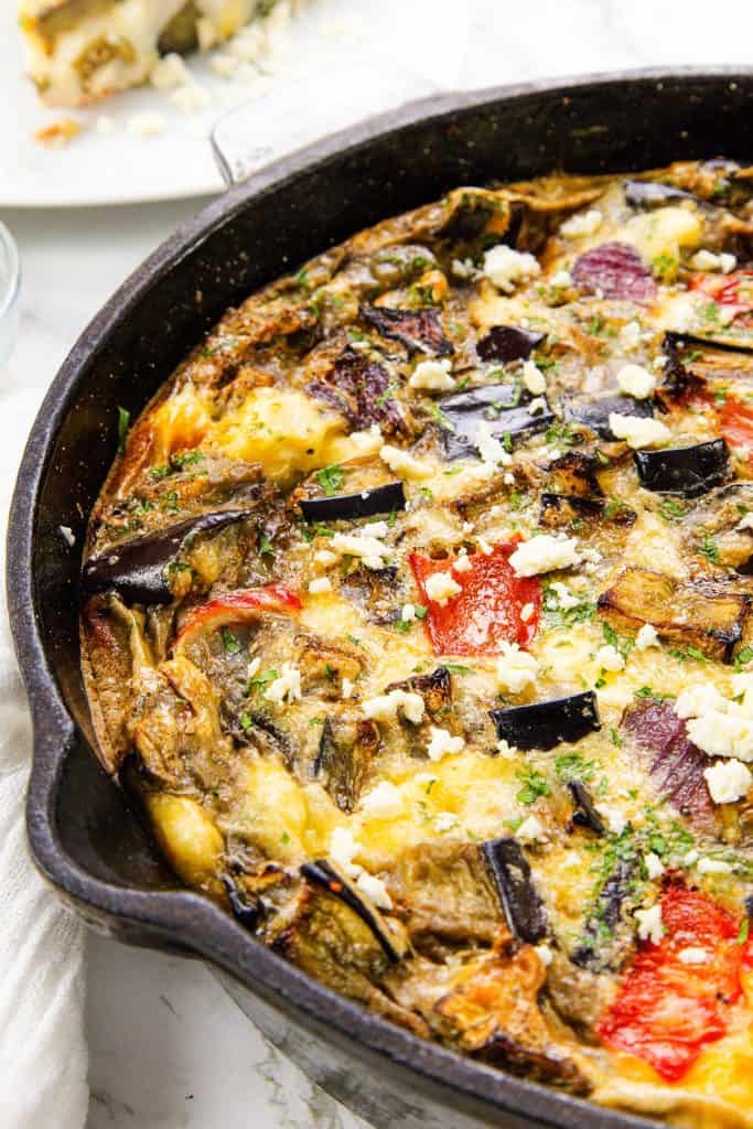 A close-up of a Baked Eggplant Frittata with Red Onion, Peppers & Feta in a black cast iron skillet, topped with hearty vegetables and crumbled cheese. A slice of this vibrant frittata is served on a plate in the background.