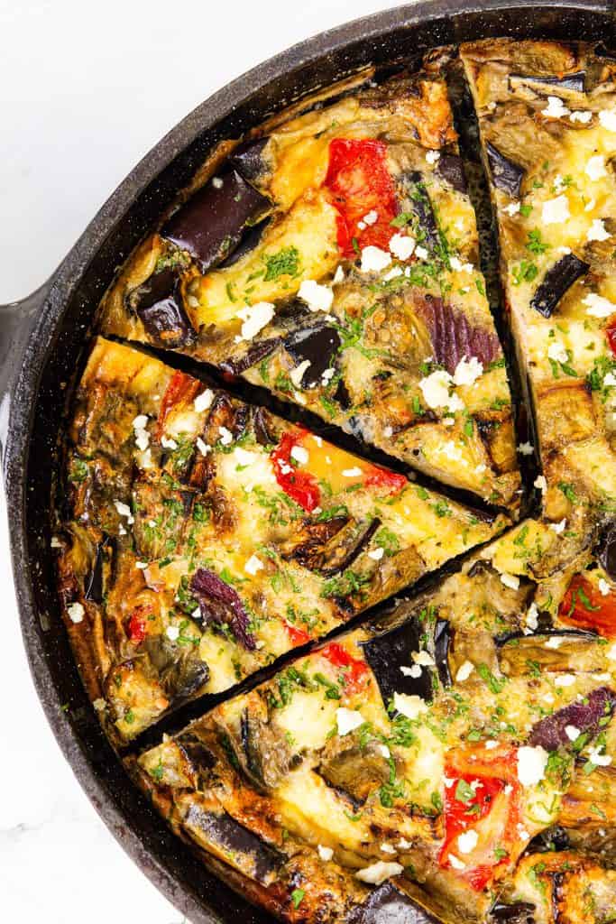 A close-up of a Baked Eggplant Frittata with Red Onion in a black skillet, cut into wedges. The frittata features visible pieces of peppers & feta, eggplant, and is garnished with herbs and crumbled cheese.