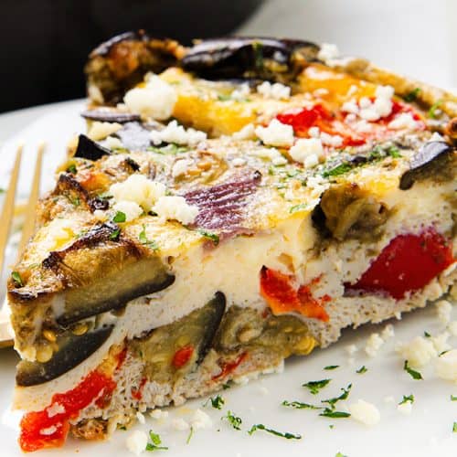A close-up of a slice of Baked Eggplant Frittata with Red Onion, Peppers & Feta, featuring visible eggplant, red bell pepper, and crumbled cheese on top, served on a white plate with two gold forks.