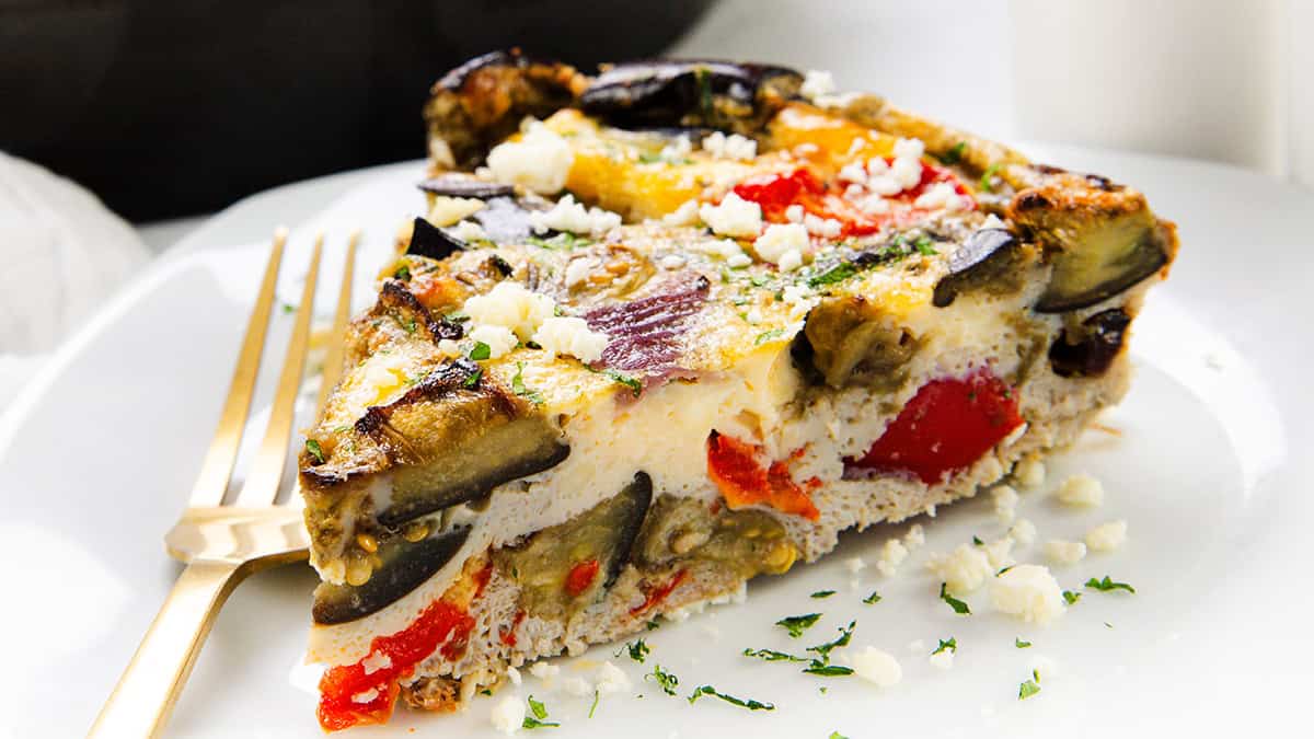 A close-up of a slice of Baked Eggplant Frittata with Red Onion, Peppers & Feta, featuring visible eggplant, red bell pepper, and crumbled cheese on top, served on a white plate with two gold forks.