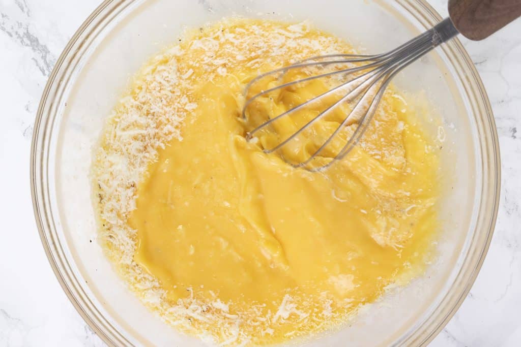 A metal whisk mixing yellow beaten eggs, grated cheese, and crumbled feta in a clear glass bowl on a white marble surface—perfect for a Peppers & Feta or Baked Eggplant Frittata with Red Onion.