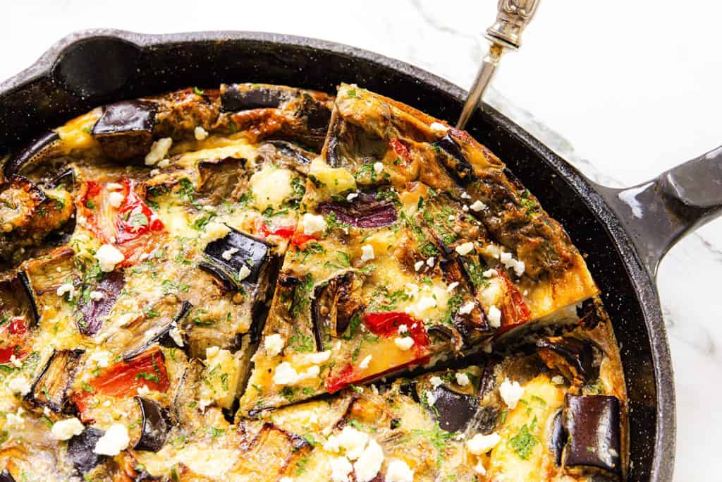 A close-up of a Baked Eggplant Frittata with Red Onion, Peppers & Feta in a black cast iron skillet, featuring visible pieces of eggplant, red peppers, herbs, and crumbled cheese, with a slice being lifted by a spatula.