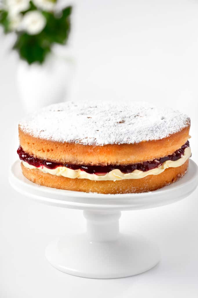 a Victoria sponge cake topped with powdered sugar on a cake stand with flowers in the background.