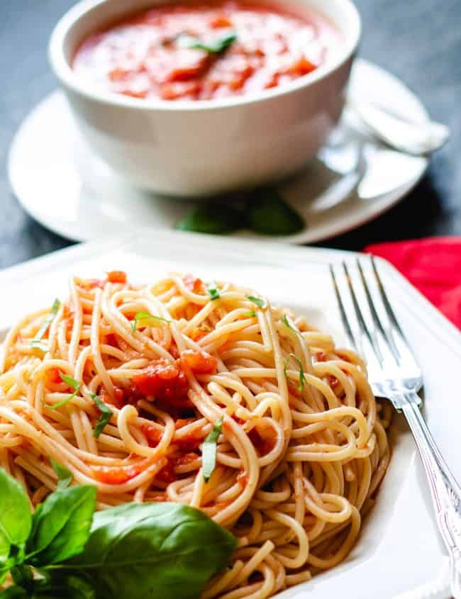 A plate of spaghetti with tomato sauce with a fork next to it and fresh basil near the dish