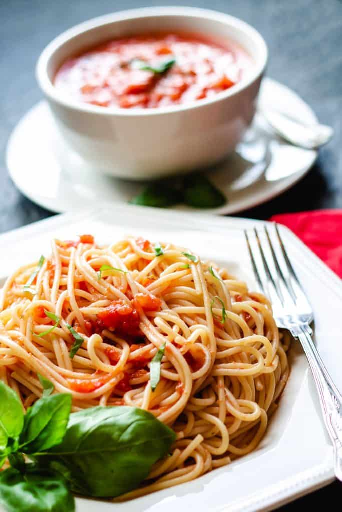 A plate of spaghetti with tomato sauce with a fork next to it and fresh basil near the dish