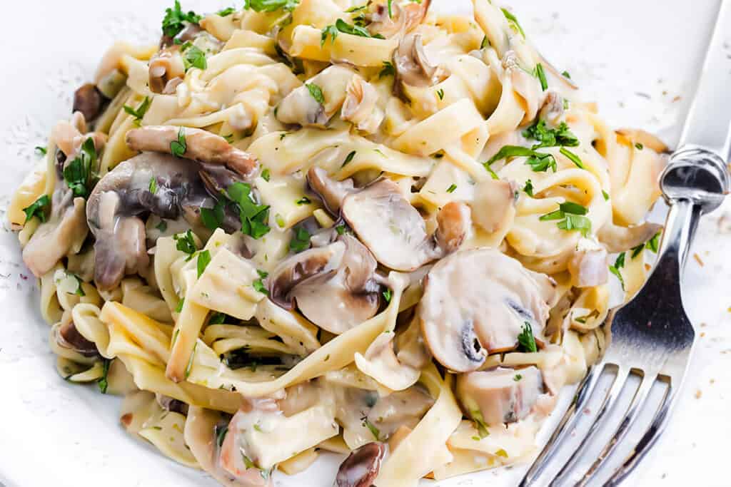 A close-up photo of a white plate overflowing with creamy tagliatelle pasta and sliced cremini mushrooms. A silver fork rests on the side of the plate.