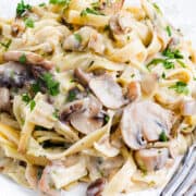 A close-up photo of a white plate overflowing with creamy tagliatelle pasta and sliced cremini mushrooms. A silver fork rests on the side of the plate.