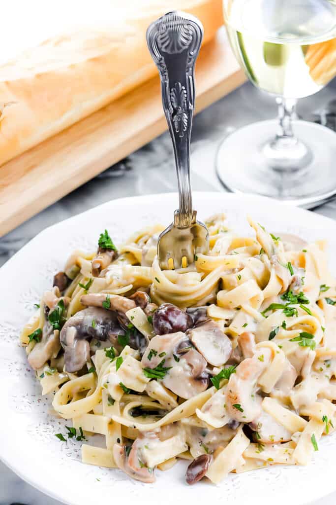 A close-up photo of a white plate overflowing with creamy tagliatelle pasta and sliced button mushrooms. A silver fork rests on the side of the plate.