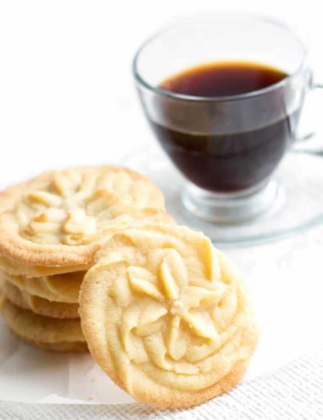 One beautiful Vanilla Shortbread Cookie sitting up against a stack of more cookies with coffee on the background.