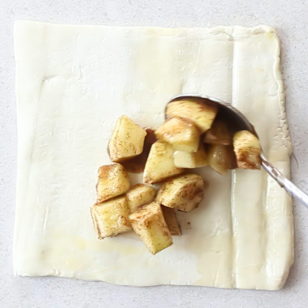 The apple mixture being spooned onto a square of puff pastry for the apple turnovers.