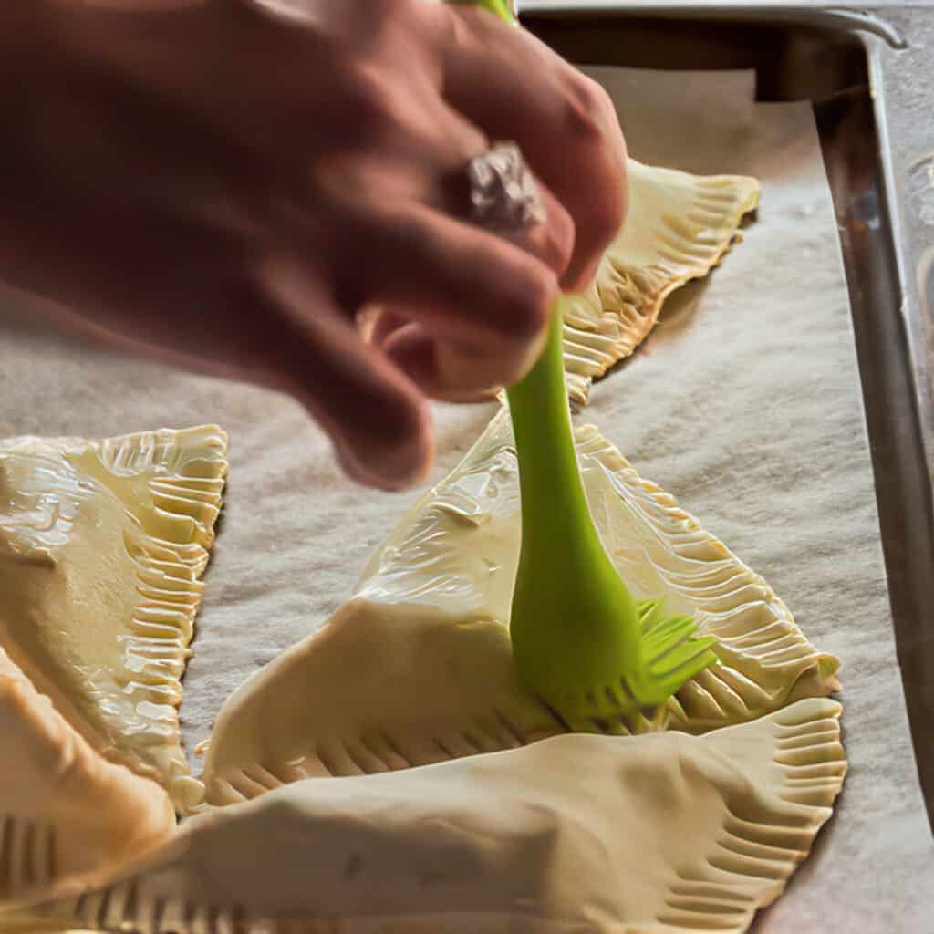 egg wash being brushed over the turnovers on the pan.