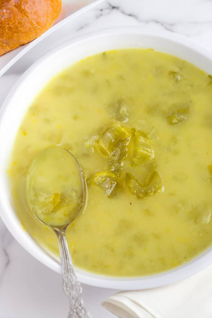 A close-up of a bowl of chunky celery soup with a spoon, showing the texture of the soup with pieces of celery visible, accompanied by a crusty bread roll on the side.