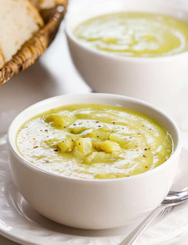 A bowl of creamy celery soup garnished with celery chunks, freshly ground black pepper, served on a white plate with a basket of bread in the background.