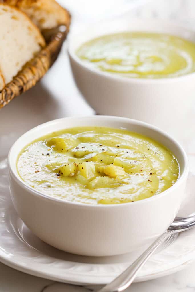 A bowl of creamy celery soup garnished with celery chunks, freshly ground black pepper, served on a white plate with a basket of bread in the background.