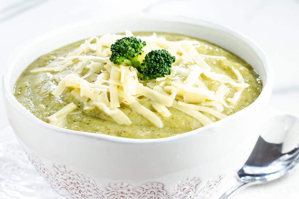 A close up image of broccoli soup in a white bowl topped with shredded cheese.