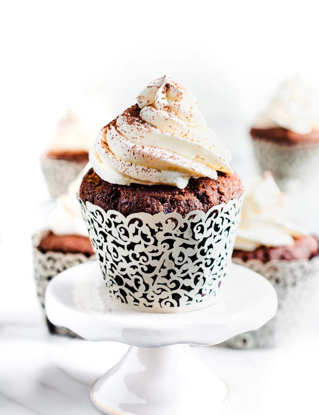 Close-up of a chocolate cupcake on a plate. The cupcake is frosted with whipped cream and dusted with cocoa powder.