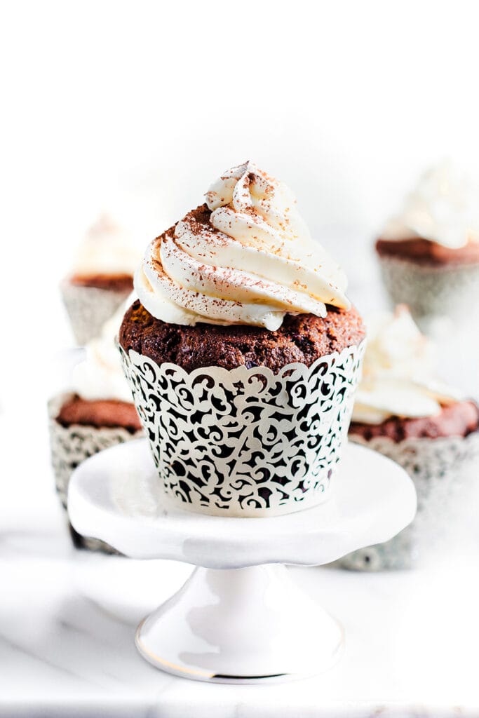 Close-up of a chocolate cupcake on a plate. The cupcake is frosted with whipped cream and dusted with cocoa powder.