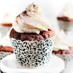 A close-up of two chocolate cupcakes on a white plate. The cupcakes are topped with whipped cream frosting and dusted with cocoa powder.