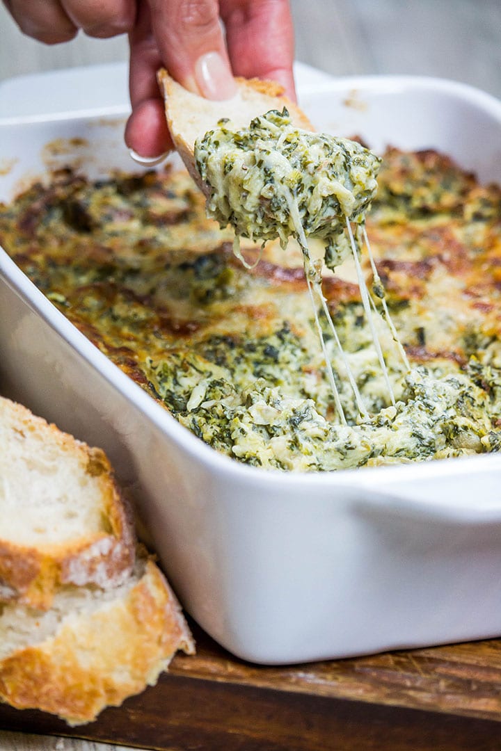 A piece of bread coming out of the dip with a string of cheese stretching from the dish