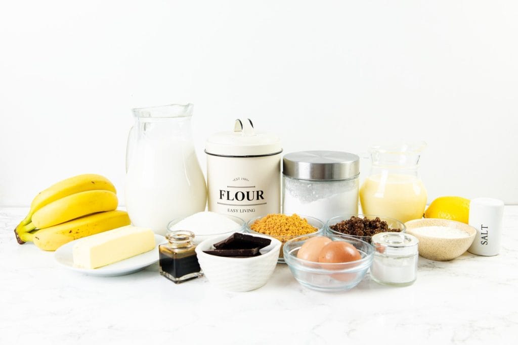 A selection of baking ingredients on a counter, including bananas, butter, eggs, flour, sugar, milk, vanilla, chocolate, brown sugar, salt, and small bowls with additional ingredients—everything you need to create a decadent Caramel Banana Cake with Whipped Cream Frosting.