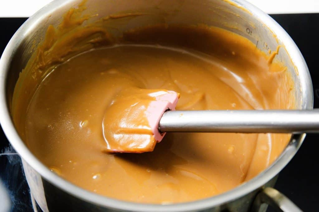 A saucepan filled with thick, smooth caramel for a Caramel Banana Cake with Whipped Cream Frosting is being stirred gently with a pink spatula.