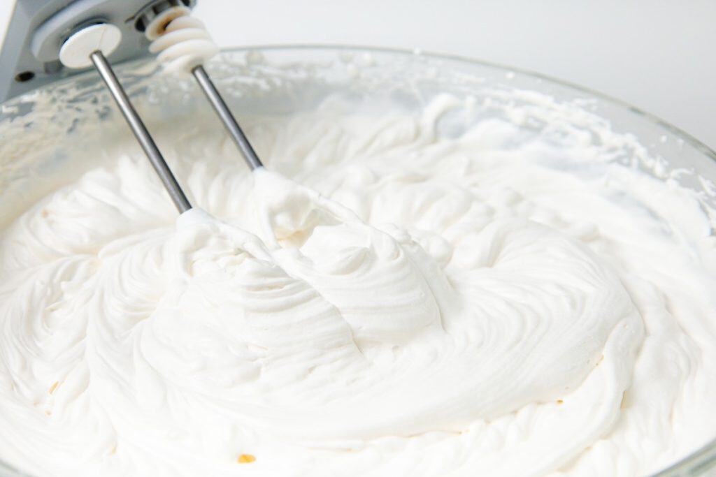 An electric mixer with beaters is whipping fluffy white cream in a glass bowl, preparing the perfect whipped cream frosting for a delicious Caramel Banana Cake.