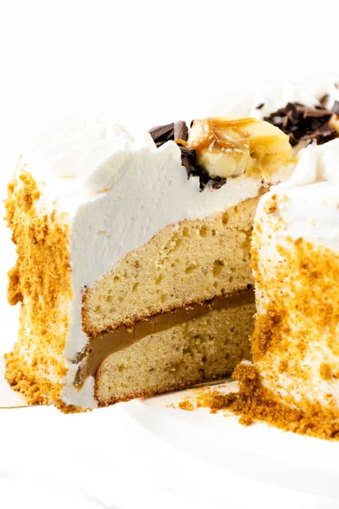 A sliced caramel banana cake with two layers, filled with a rich caramel or coffee-flavored cream, topped with luscious whipped cream frosting, caramel drizzle, chocolate shavings, and surrounded by crumbled cookie or cake bits.