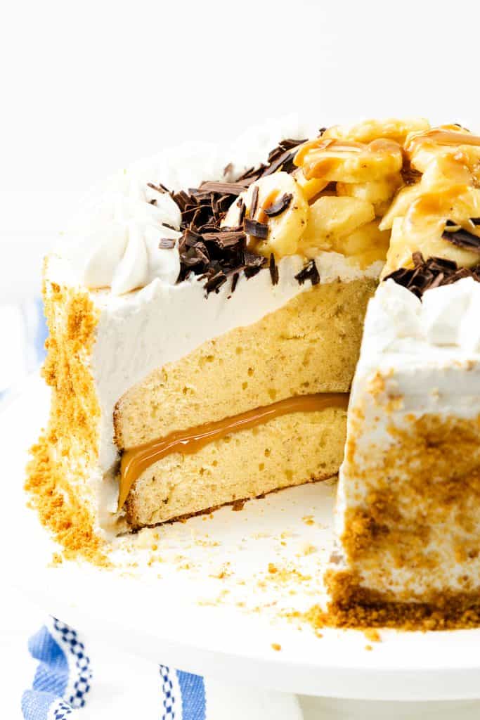 A Caramel Banana Cake with Whipped Cream Frosting, this decadent layered cake features smooth white frosting, luscious caramel filling, chocolate shavings, fresh banana slices, and a rich caramel drizzle. One large slice is missing, revealing the moist interior. The cake sits on a white plate with crumbs scattered around the edge.