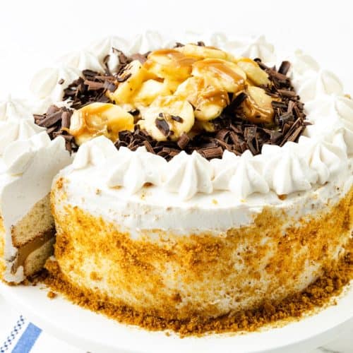 A decadent Caramel Banana Cake with Whipped Cream Frosting is topped with fluffy cream, fresh banana slices, chocolate shavings, and a luscious drizzle of caramel sauce. Sitting on a white plate, this cake reveals its layered interior and crumb-coated edge as a slice is being lifted out.
