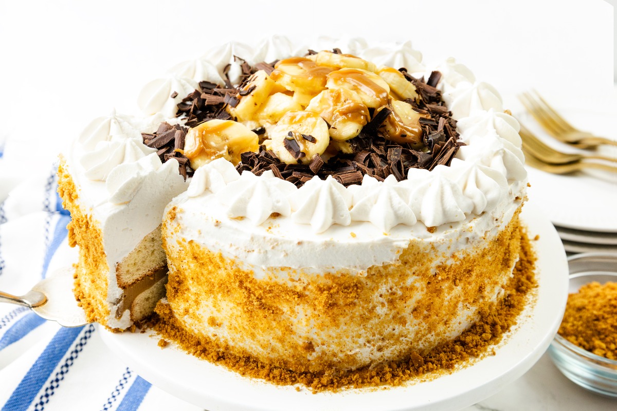 A decadent Caramel Banana Cake with Whipped Cream Frosting is topped with fluffy cream, fresh banana slices, chocolate shavings, and a luscious drizzle of caramel sauce. Sitting on a white plate, this cake reveals its layered interior and crumb-coated edge as a slice is being lifted out.