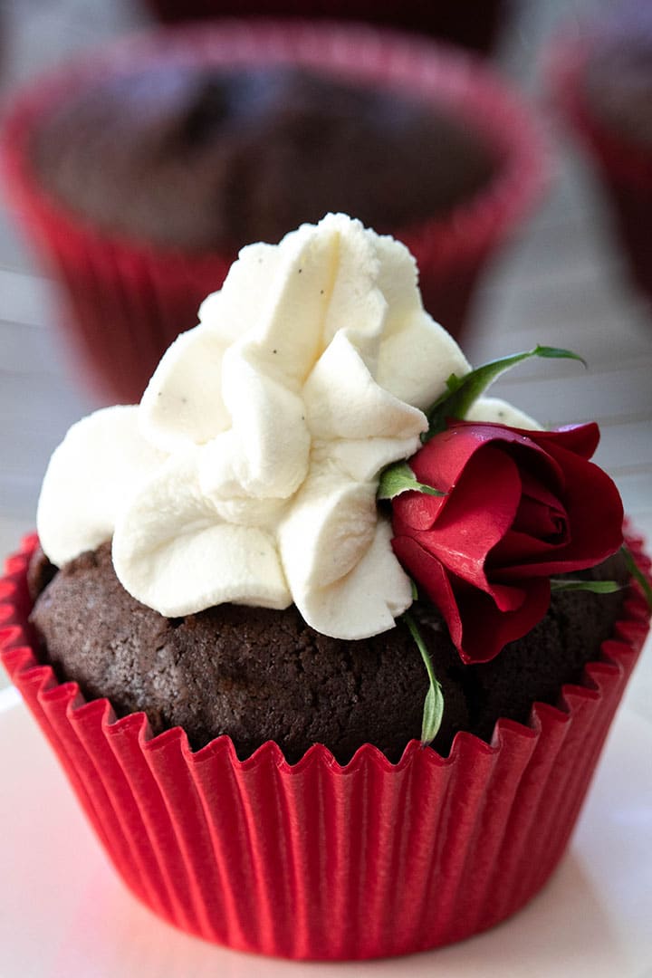 A chocolate cupcake with a red casing, whipped cream icing and a small red rose for decoration